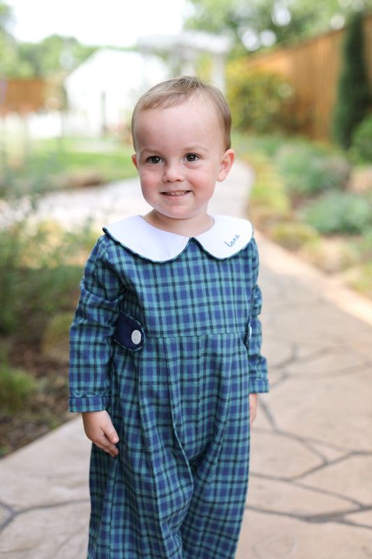 young boy wearing a blue and green plaid long romper with a white collar outdoors.