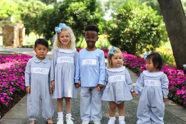 Five children wearing personalized light blue custom smocked outfits with name panels outdoors