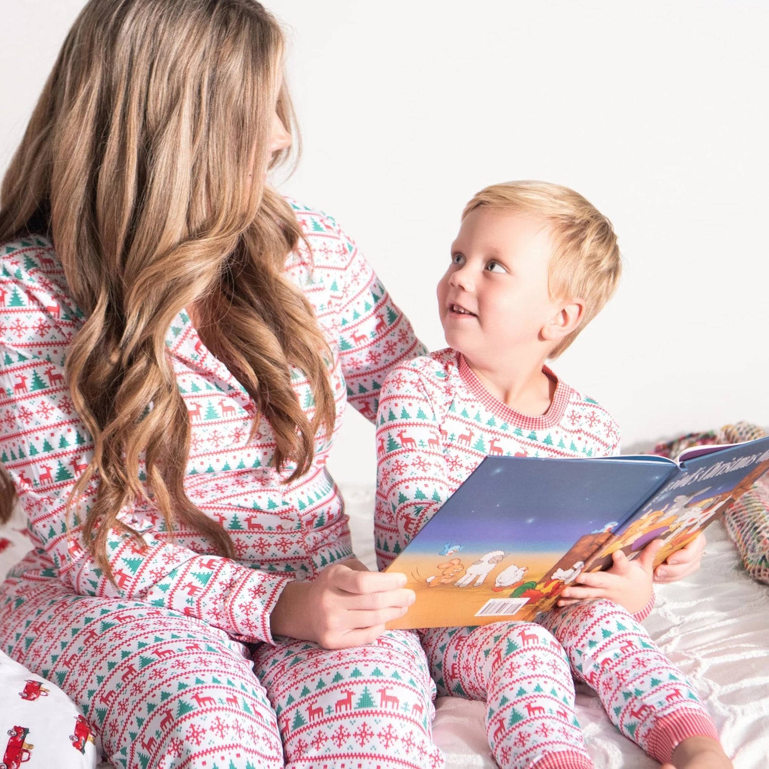A woman and young boy sit together in matching Christmas pajamas with red, green, and white patterns while reading a children’s book on a bed.