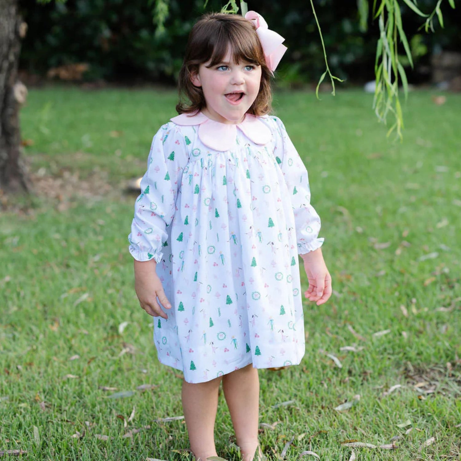 A young girl stands outdoors wearing a long-sleeve white Christmas dress with pastel holiday prints and a pink Peter Pan collar, smiling beneath tree branches.