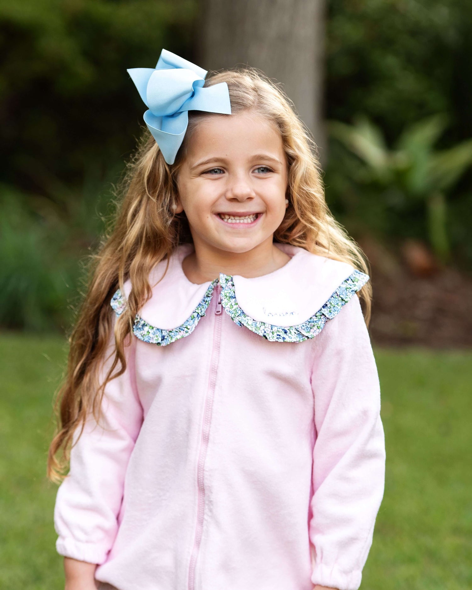 Young girl wearing a light pink jacket with a floral Peter Pan collar and a large blue bow, standing outdoors.