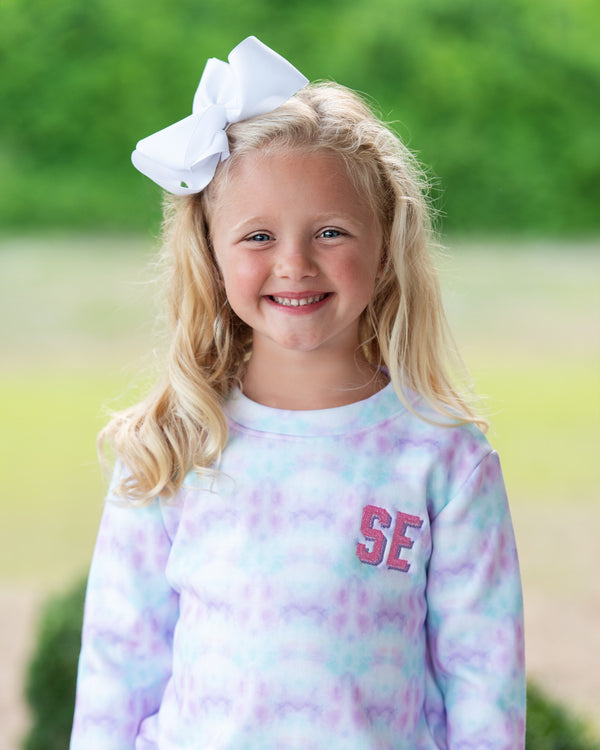 Girl wearing a pastel tie-dye long sleeve top with pink SE lettering and a large white bow, standing outdoors.