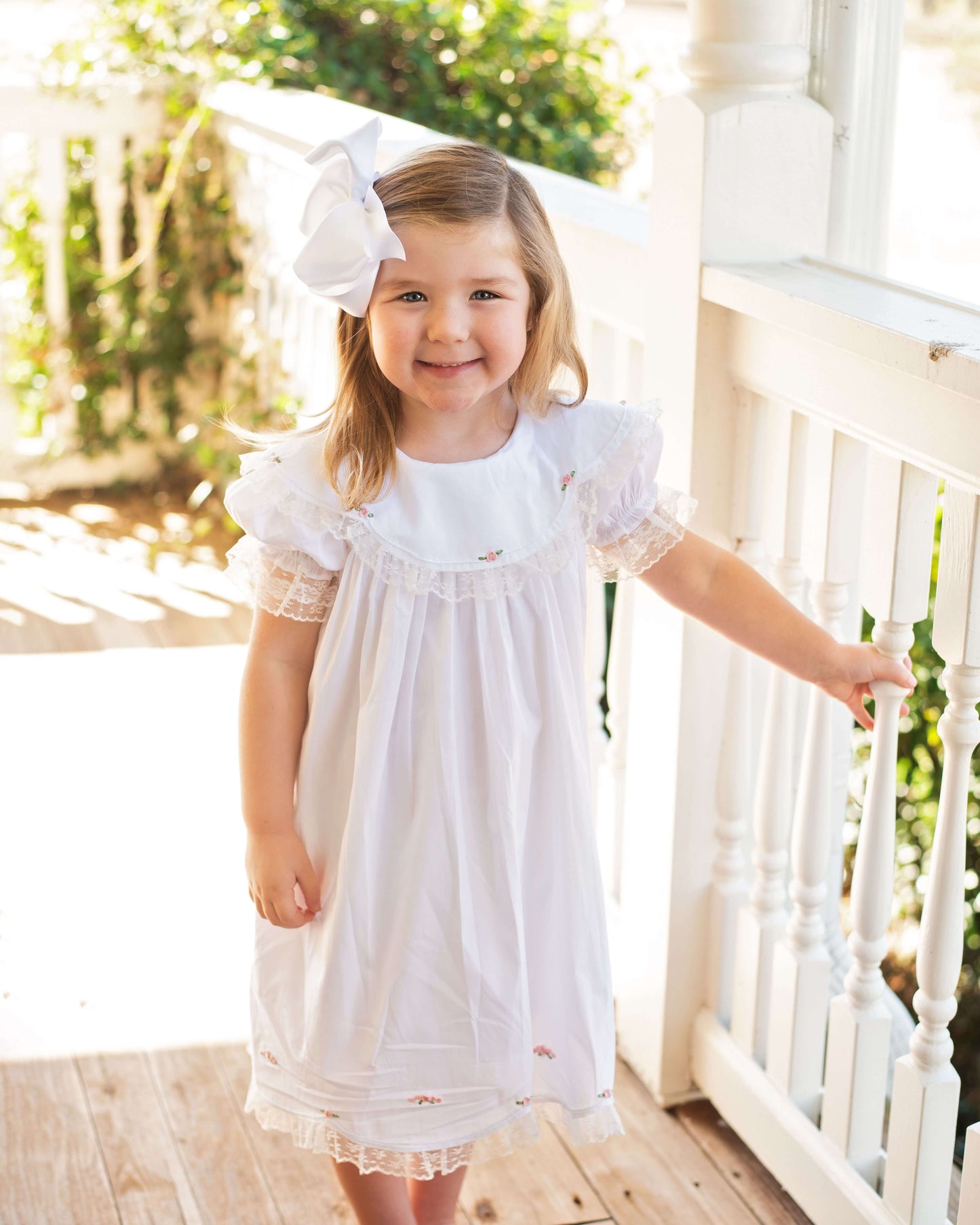 Young girl in a white dress with a large bow standing on a wooden deck.