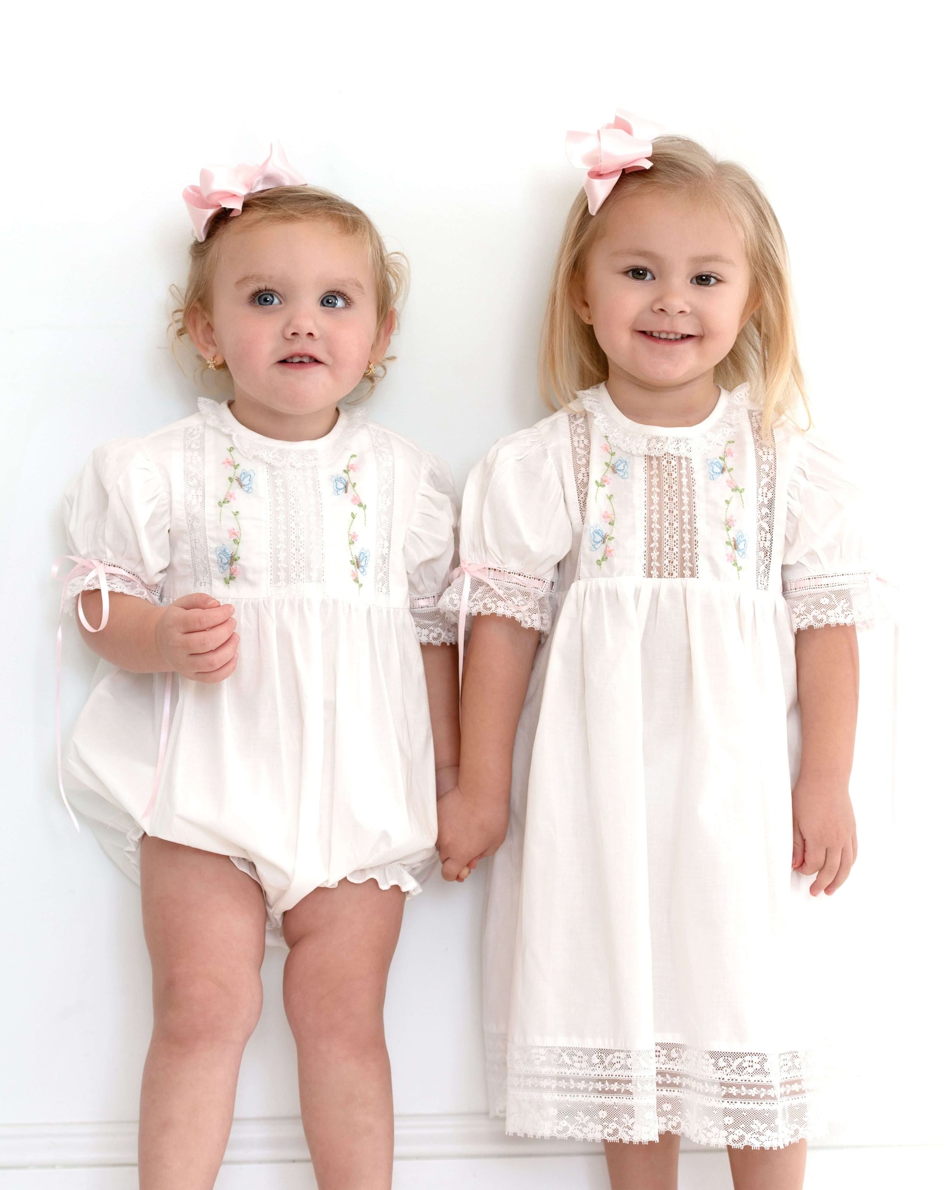 Two young girls wearing white dresses with lace details against a white background