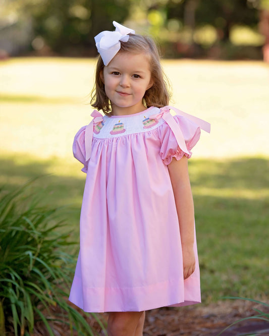Young girl wearing a pink dress with ruffled sleeves and a white bow in her hair, standing outdoors.