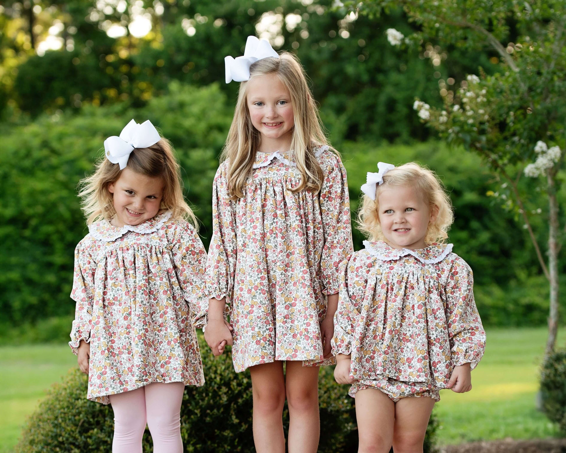 Three young girls in matching floral dresses standing outdoors with greenery in the background.