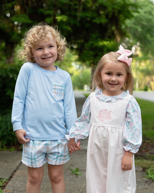 Two young children standing outdoors and holding hands. The child on the left wears a light blue long-sleeve shirt with a small pocket and matching plaid shorts.