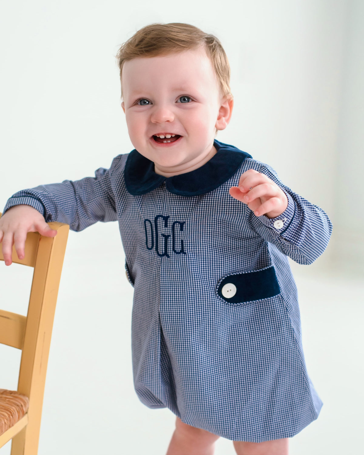 A baby boy wearing a blue and white gingham romper with a navy blue velvet collar and chest pocket, standing next to a wooden chair.