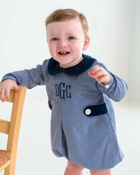 A baby boy wearing a blue and white gingham romper with a navy blue velvet collar and chest pocket, standing next to a wooden chair.