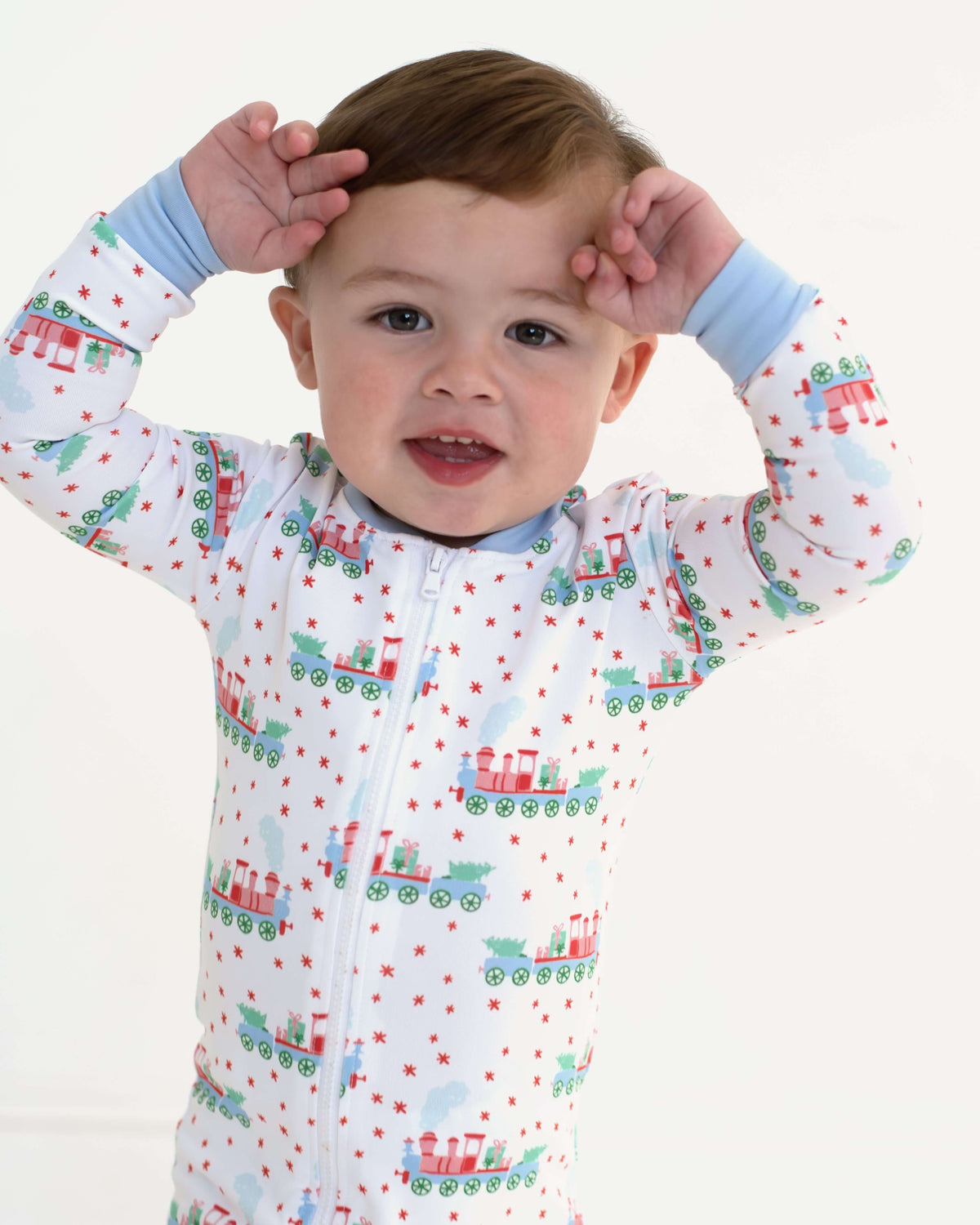 A baby wearing a white long-sleeve zip-up pajama with a colorful Christmas train pattern, posed with hands on head against a plain white background.