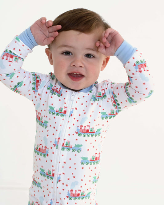 A baby wearing a white long-sleeve zip-up pajama with a colorful Christmas train pattern, posed with hands on head against a plain white background.
