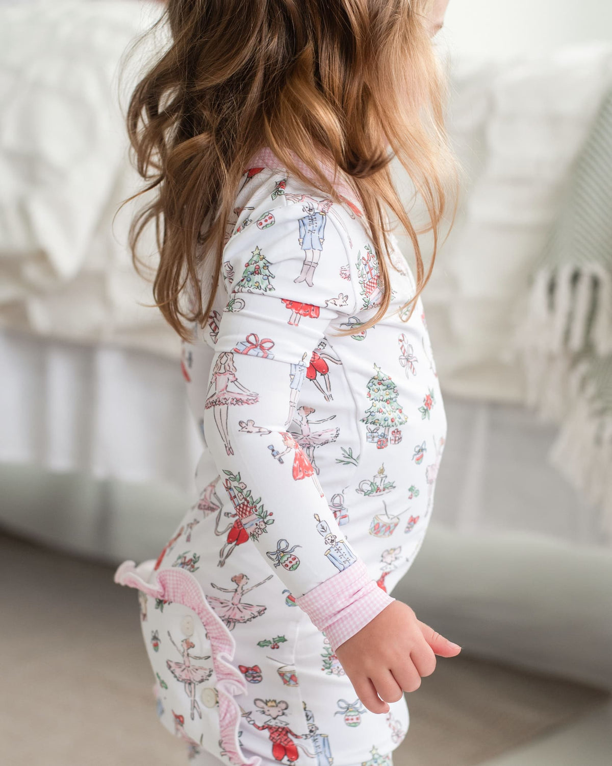 A child wearing a white long-sleeve pajama top with a colorful ballet dancer and holiday print, shown from the side in a softly lit bedroom setting.