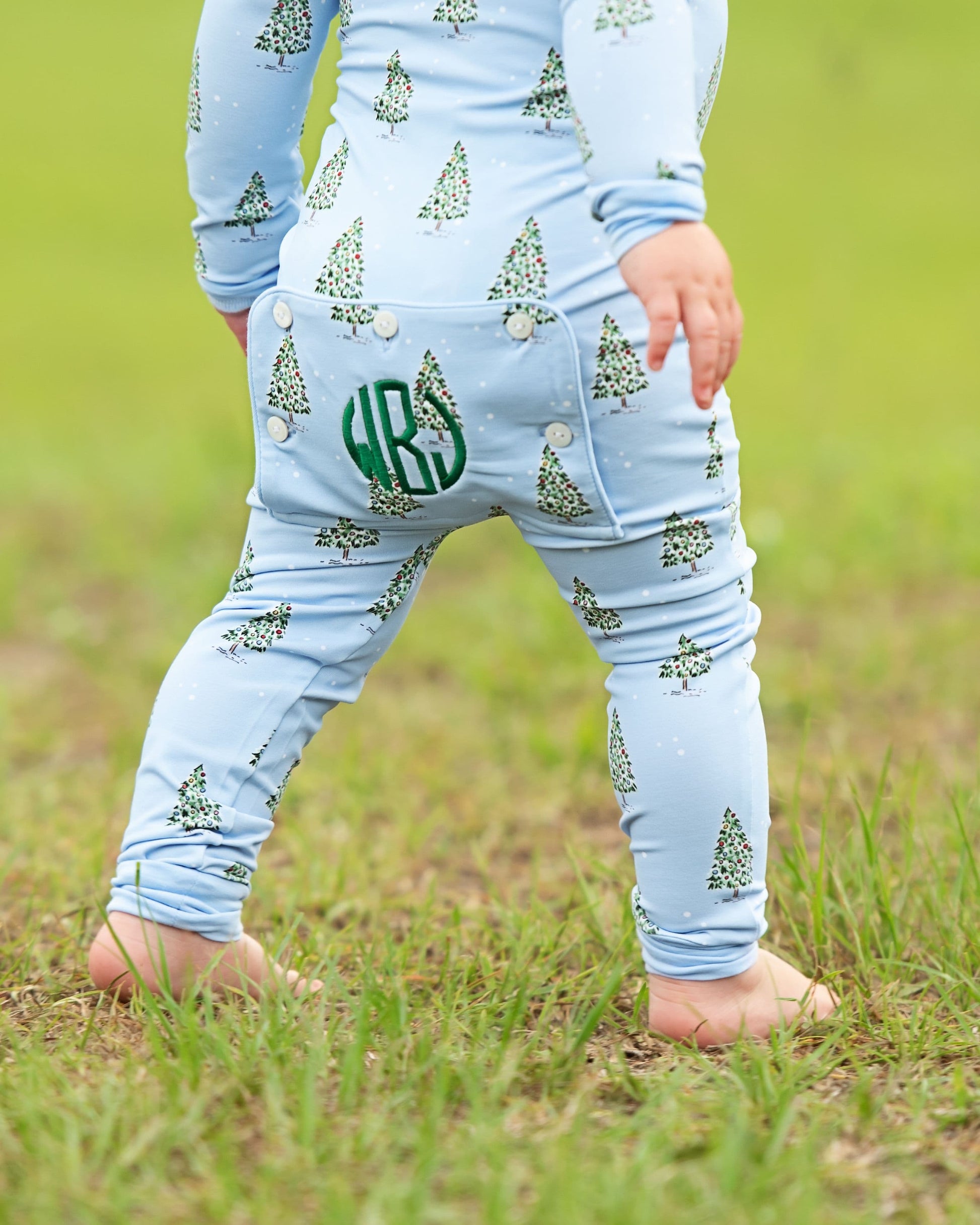 A close-up back view of a child wearing a light blue long-sleeve one-piece outfit with a green Christmas tree print and a green monogram patch on the back, standing barefoot on grass.