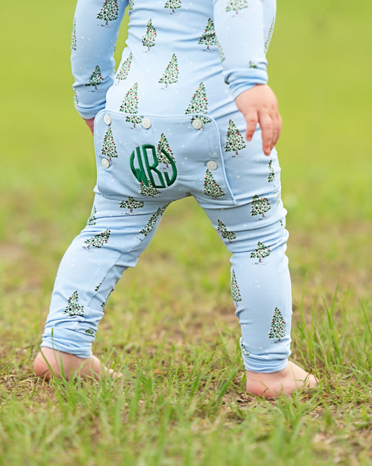 A close-up back view of a child wearing a light blue long-sleeve one-piece outfit with a green Christmas tree print and a green monogram patch on the back, standing barefoot on grass.