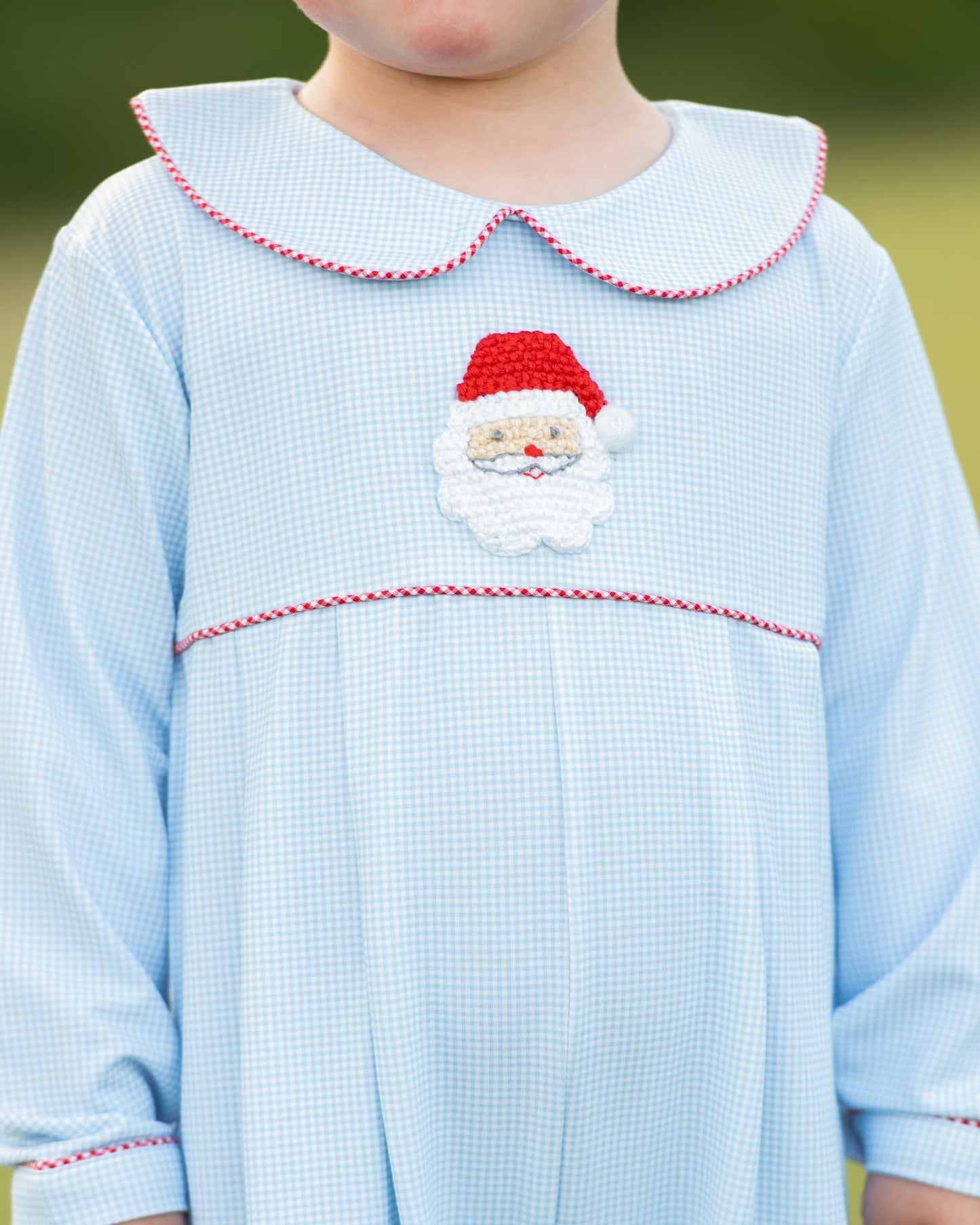 A close-up of a child wearing a light blue gingham dress with a red and white braided Peter Pan collar, a red and white braided horizontal trim line, and a central Santa Claus face applique.