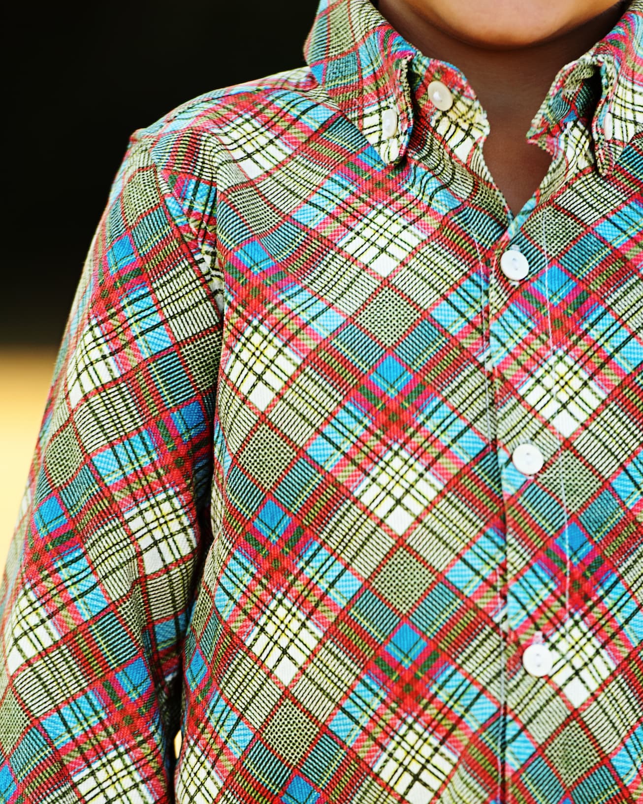 A close-up of a child wearing a long-sleeve button-up shirt with a vibrant multicolor plaid pattern featuring red, blue, green, and beige stripes.