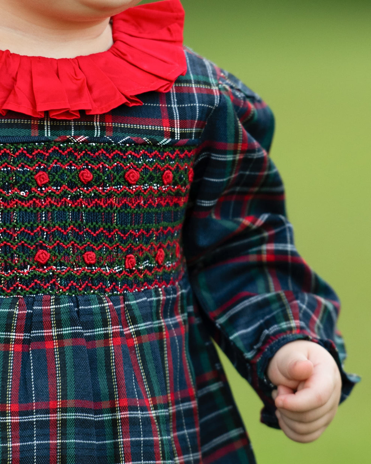 A close-up of a child wearing a long-sleeve plaid dress with a red ruffled collar and red embroidered smocked bodice details, set against a soft green blurred background.