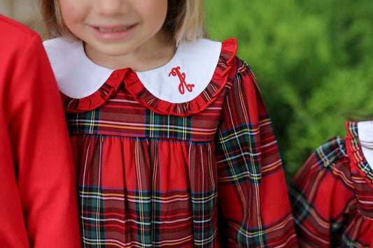 A close-up of a child wearing a red plaid dress with a white ruffled Peter Pan collar and red embroidered initials on the collar, set against a blurred green outdoor background.