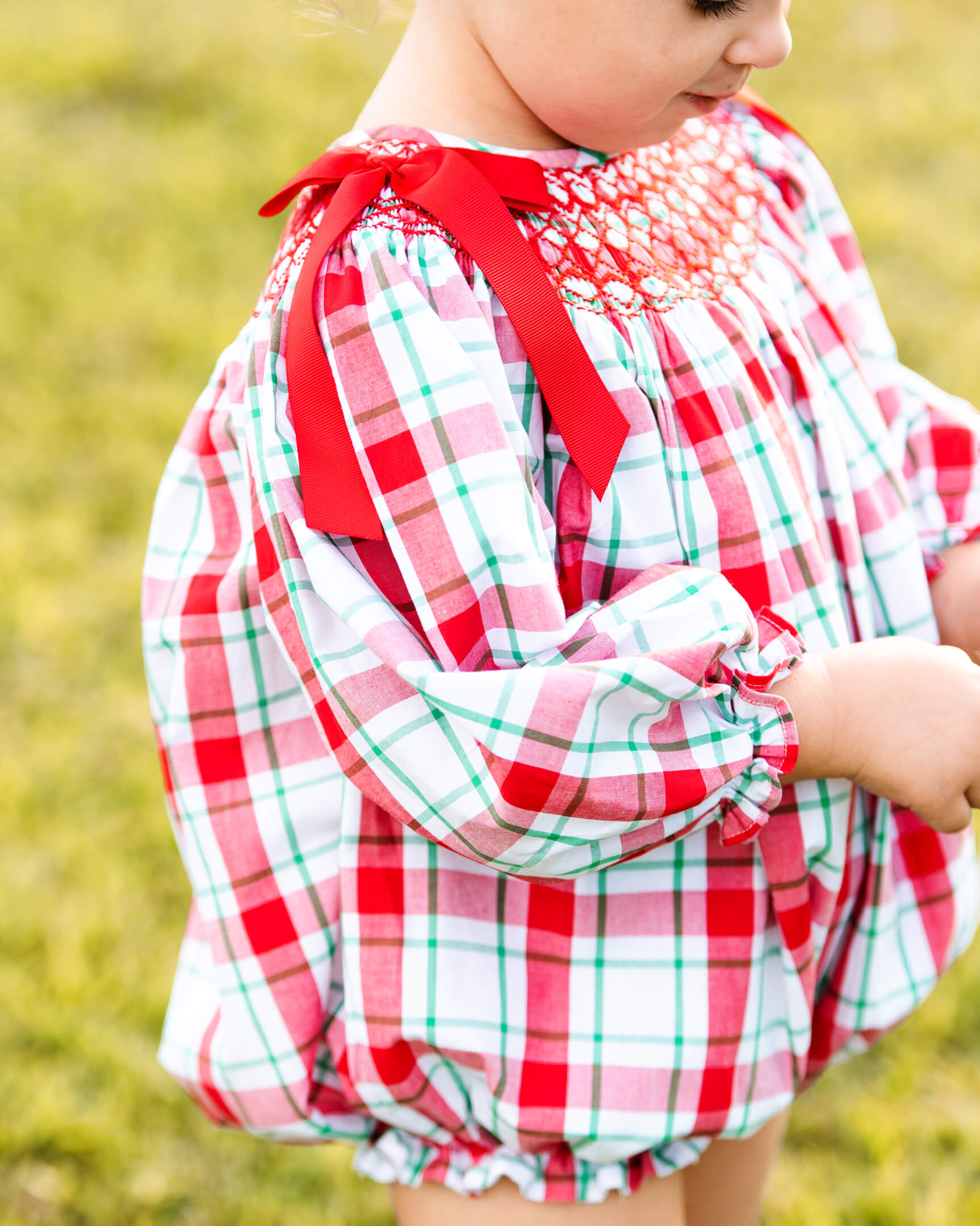 A close-up of a child wearing a red, white, and green plaid long-sleeve dress with a smocked chest and red ribbon bows, photographed outdoors on a grassy background.