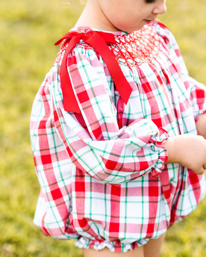 A close-up of a child wearing a red, white, and green plaid long-sleeve dress with a smocked chest and red ribbon bows, photographed outdoors on a grassy background.