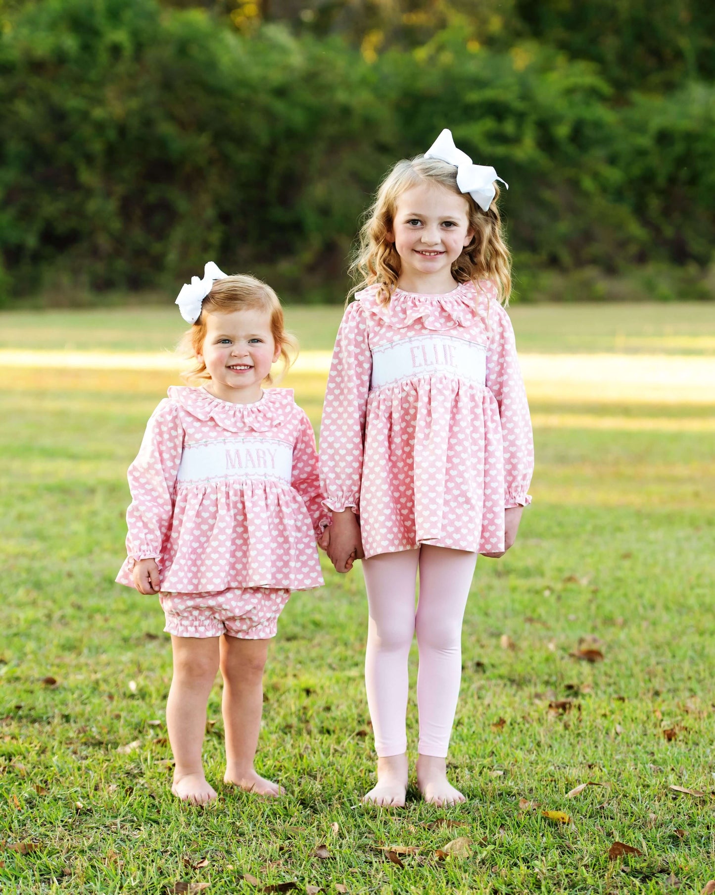 A couple of young girls in matching pink heart outfits standing on grass with a blurred background