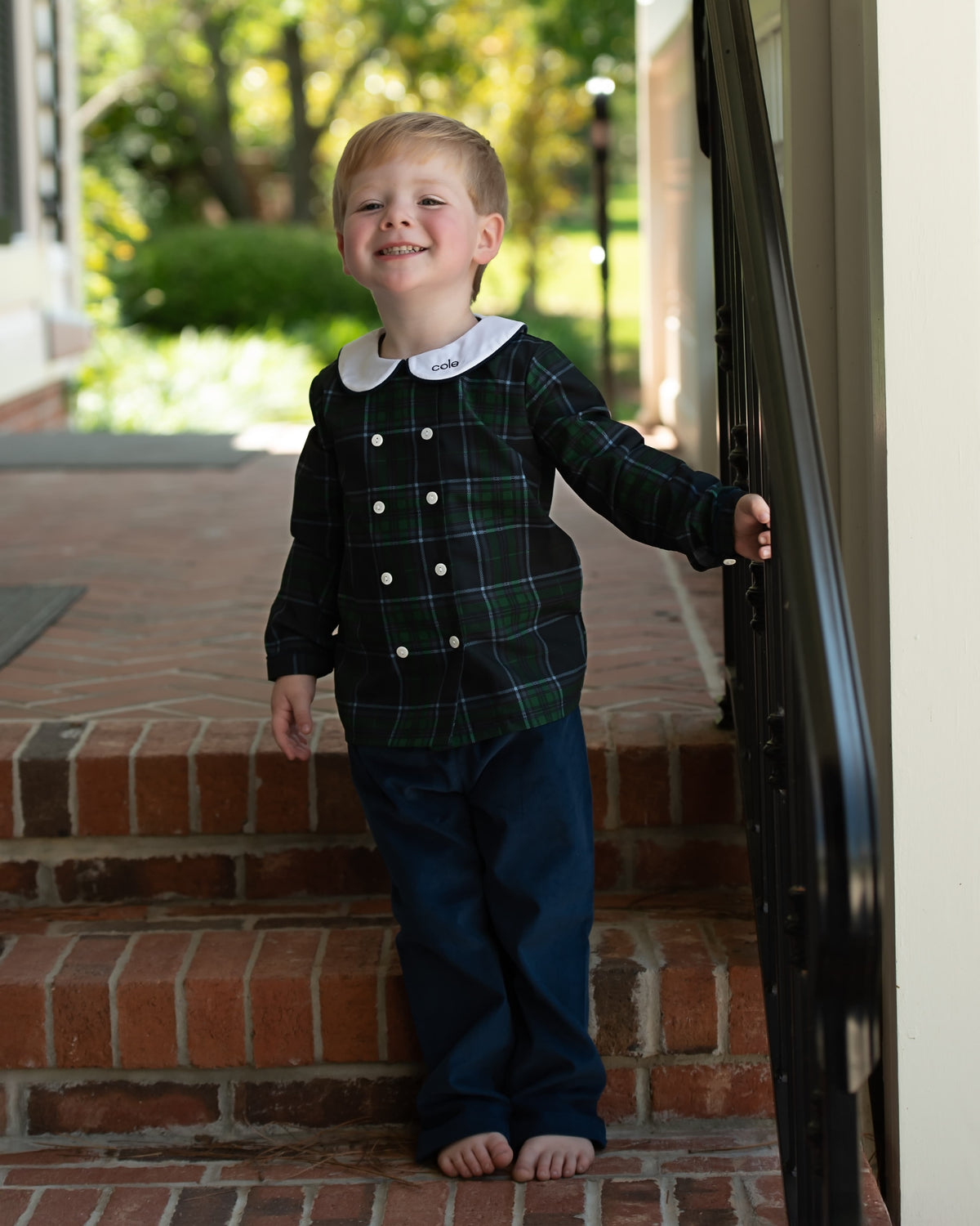 A little boy standing outside wearing a navy and green plaid peter pan pant set with a small monogram on the collar