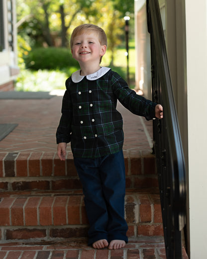 A little boy standing outside wearing a navy and green plaid peter pan pant set with a small monogram on the collar