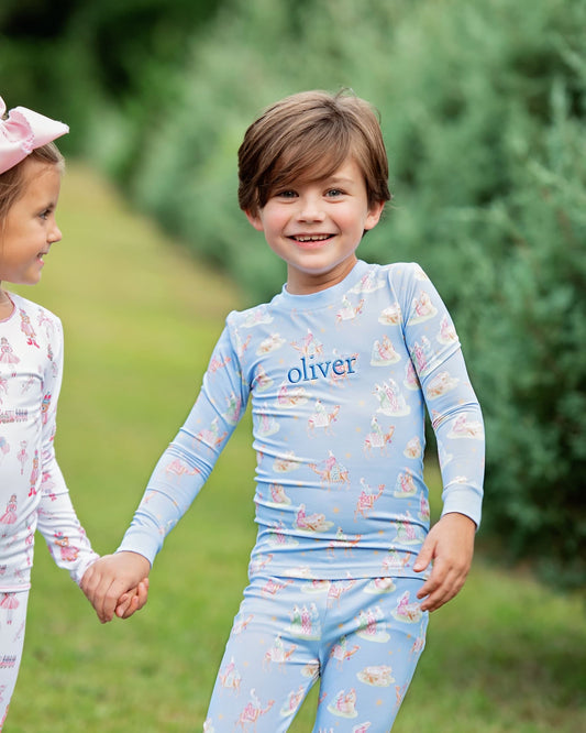 A smiling young boy wearing a light blue long-sleeve pajama set with a bunny print and the name 'oliver' on the chest, standing outdoors in a grassy area and holding hands with a girl in a matching pajama set.