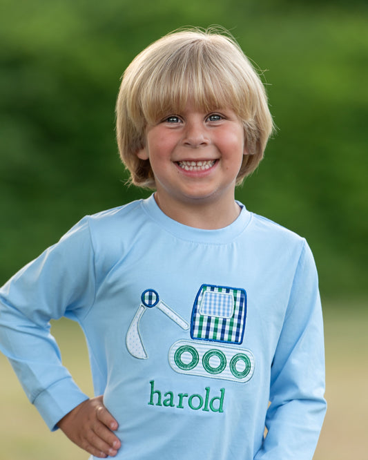 A smiling young boy with blonde hair wearing a light blue long-sleeve shirt with a green and blue gingham construction truck graphic and the name 'harold' embroidered in green, set against a soft-focus green outdoor background.