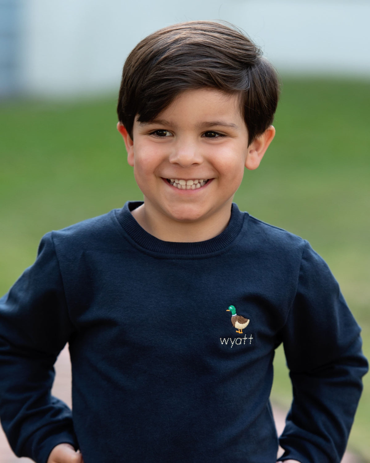 A smiling young boy with dark brown hair wearing a navy blue long-sleeve crewneck sweatshirt with a small embroidered rooster and the name 'wyatt' on the left chest, set against a soft-focus green outdoor background.