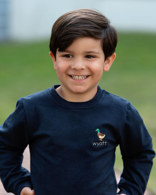 A smiling young boy with dark brown hair wearing a navy blue long-sleeve crewneck sweatshirt with a small embroidered rooster and the name 'wyatt' on the left chest, set against a soft-focus green outdoor background.