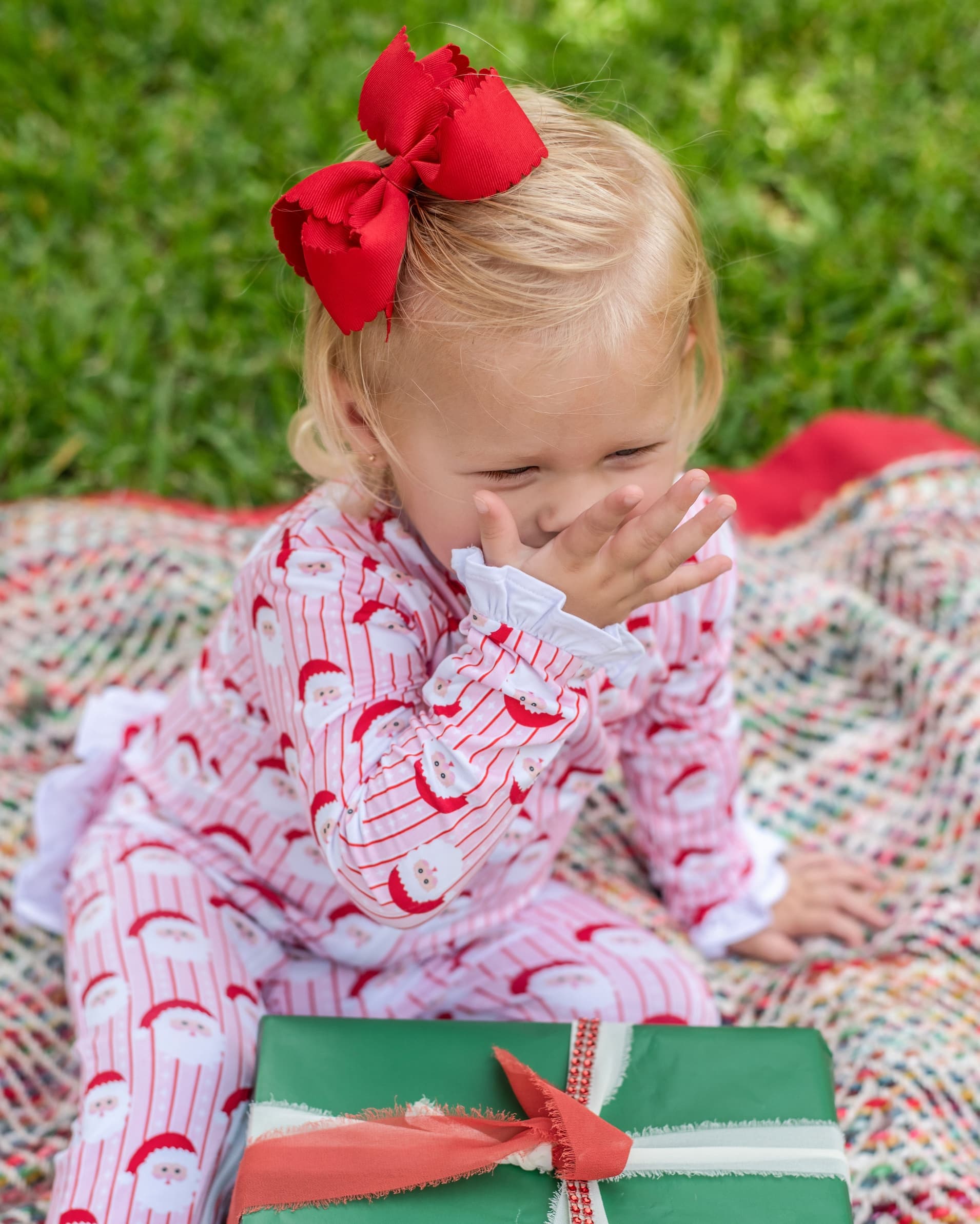A toddler girl sitting on a colorful blanket outdoors on grass, wearing a red bow in her blonde hair and a pink Santa-themed pajama romper. A green gift box with a red ribbon is in the foreground.