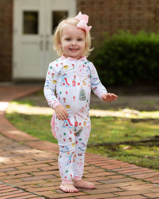 A toddler girl stands barefoot on a brick walkway outdoors, wearing a white long-sleeve footless pajama romper covered in colorful holiday illustrations, with a pink bow in her hair.