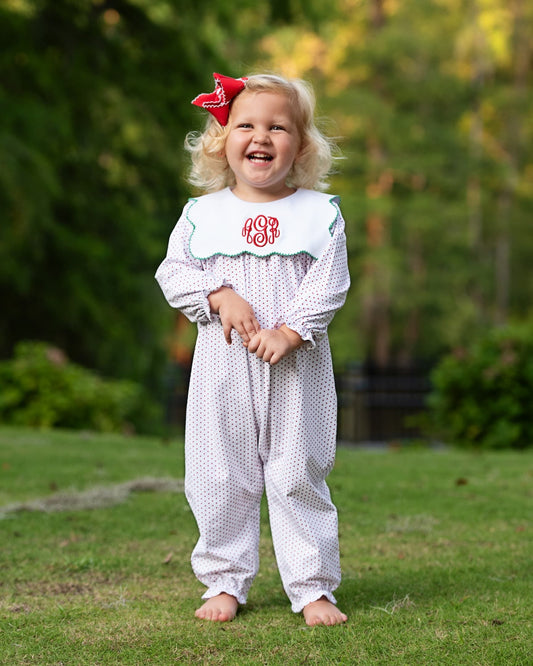 A toddler girl stands barefoot on a grassy lawn outdoors, wearing a white long-sleeve footless jumpsuit with small red polka dots and a white bib with red monogram embroidery. She has a red bow in her hair, and the background is a blurred green garden.