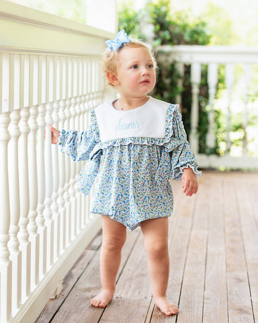 A toddler girl stands barefoot on a wooden porch next to a white railing, wearing a blue and white floral romper with a white bib collar that says 'Aleo' in blue script, and a light blue bow in her hair.