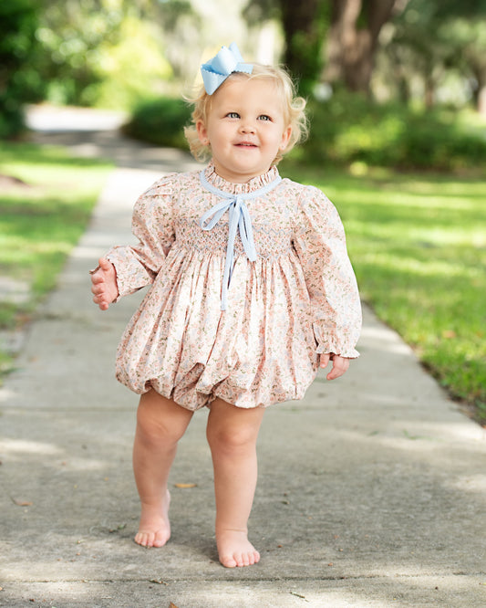 A toddler girl walking barefoot on a paved path outdoors, wearing a light pink smocked romper with a subtle floral print, a light blue bow at the neck, and a matching light blue hair bow.