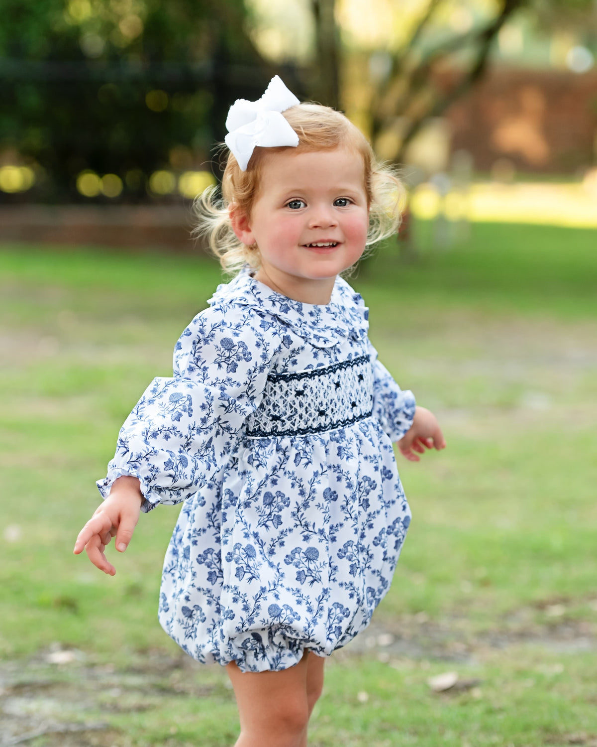 A toddler girl wearing a blue and white floral print bubble romper with smocked detailing on the chest, a white bow in her hair, and smiling outdoors on a grassy lawn.