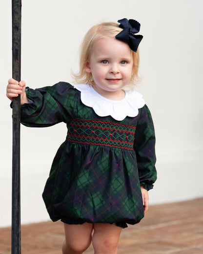 A toddler girl wearing a dark green and black plaid smocked dress with a white ruffled collar and a navy bow in her hair, holding a black metal post.