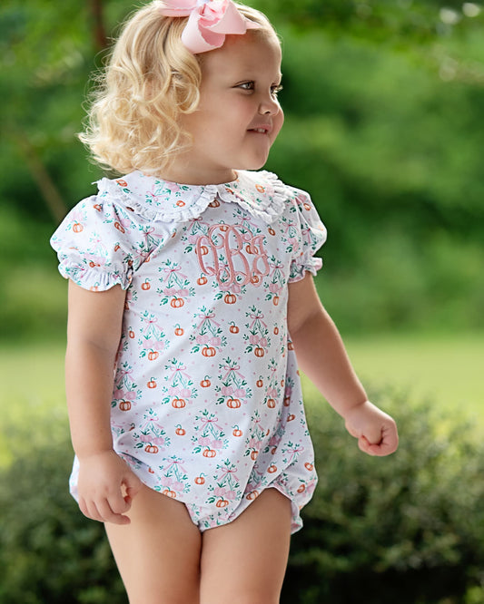 A toddler girl wearing a short-sleeve white romper with a delicate orange and green floral print and pink monogram lettering on the chest, standing outdoors in a grassy area.