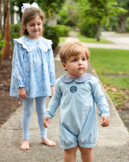 A toddler in a light blue long-sleeve romper with a navy monogram and a striped collar walks on a sidewalk, while a young girl in a matching light blue patterned dress and leggings stands behind him on the same path.
