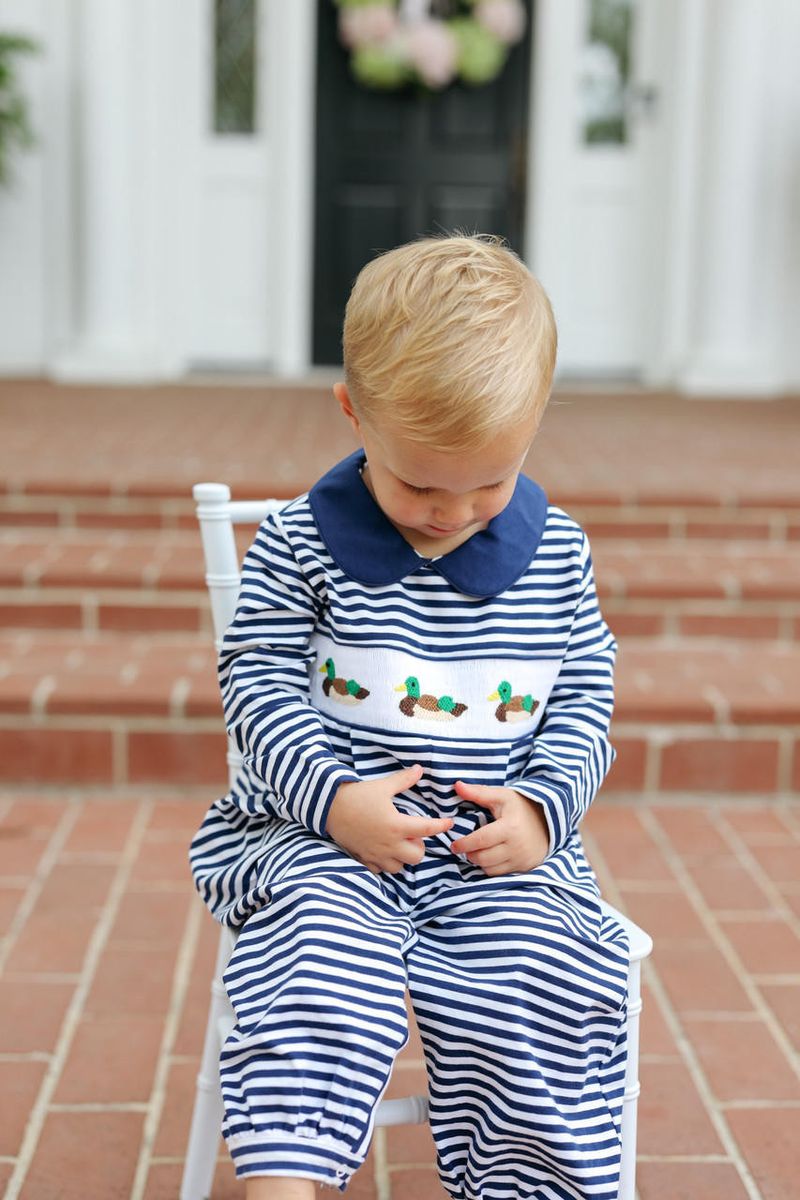 A toddler sitting on a white chair outdoors on brick steps, wearing a navy and white striped long-sleeve romper with a white collar and embroidered chest detail.