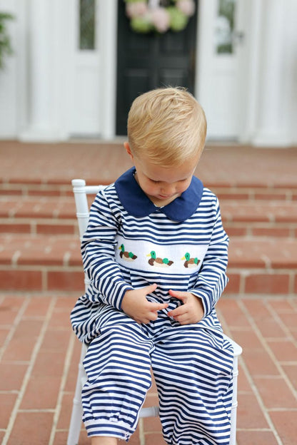 A toddler sitting on a white chair outdoors on brick steps, wearing a navy and white striped long-sleeve romper with a white collar and embroidered chest detail.