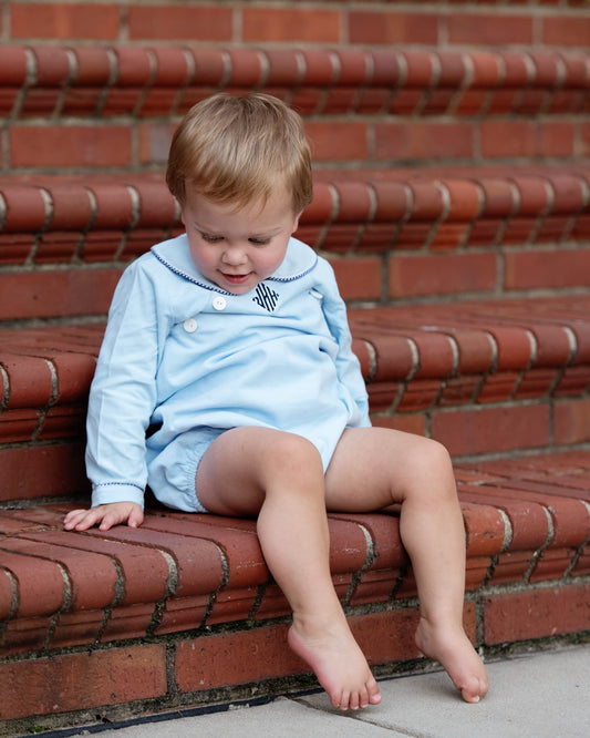 A toddler sitting on red brick steps wearing a light blue long-sleeve romper with a dark navy collar and a small embroidered monogram on the chest.