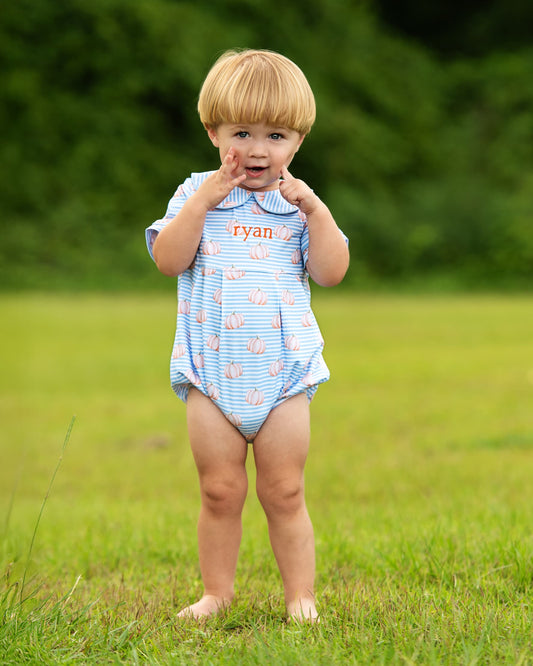 A toddler standing barefoot on a grassy field wearing a light blue and white striped short-sleeve romper with pink flamingo prints and the name 'Ryan' printed on the chest.