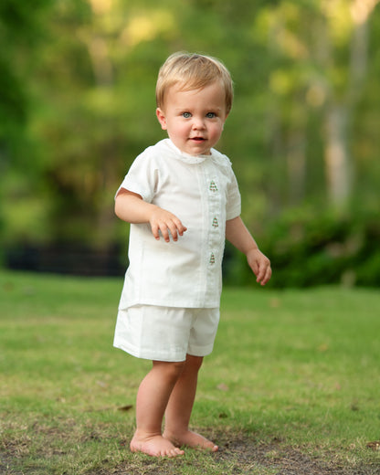 A toddler standing barefoot on a grassy lawn wearing a white short-sleeve button-front top and matching shorts, photographed outdoors with a blurred green background.