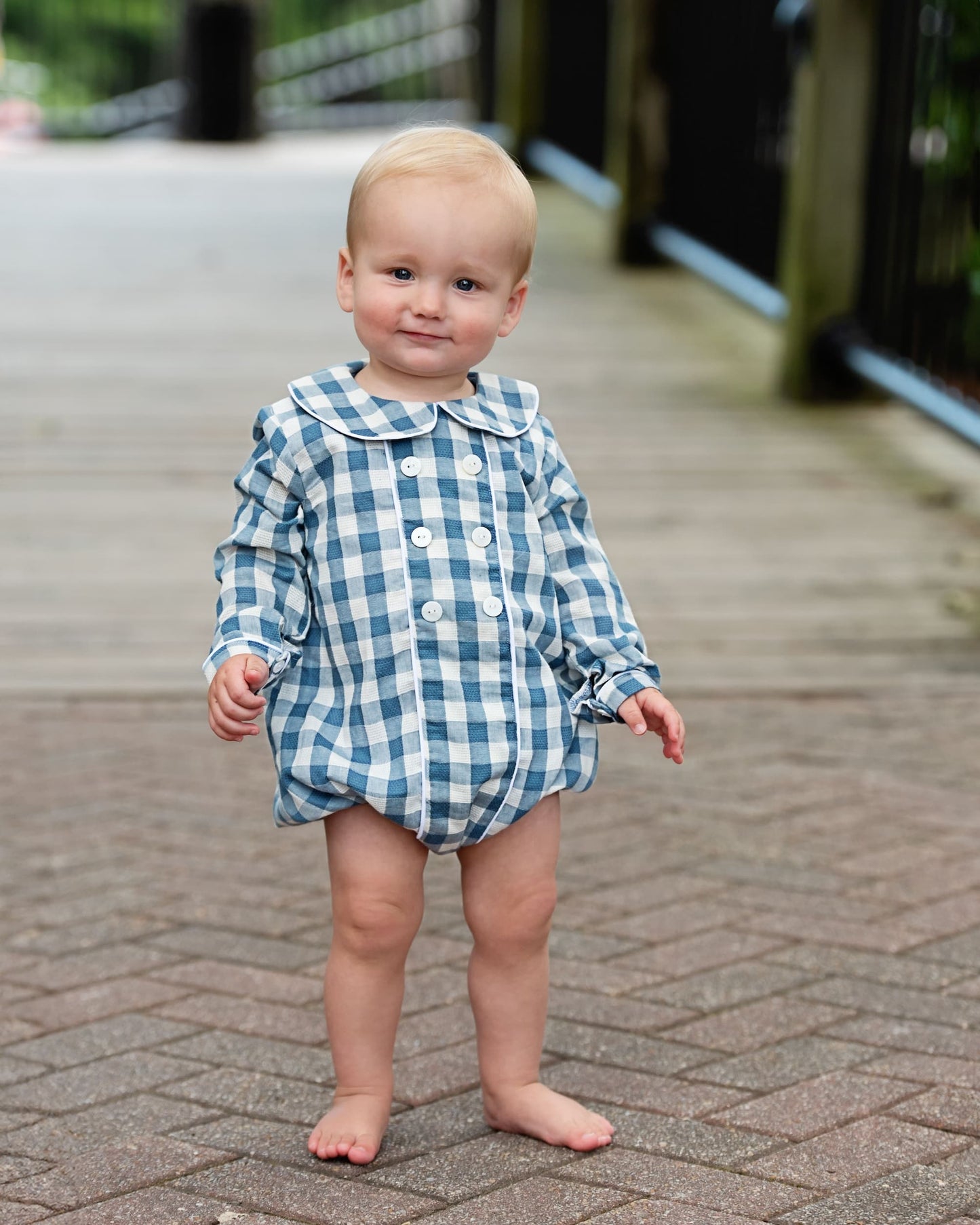 A toddler standing barefoot on a paved outdoor walkway, wearing a blue and white gingham long-sleeve romper with a button front and large collar.