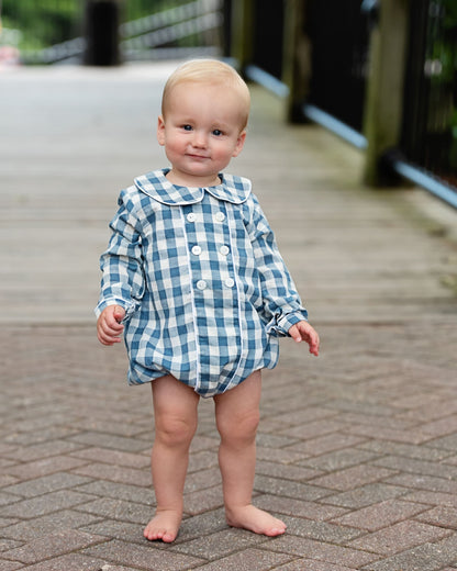 A toddler standing barefoot on a paved outdoor walkway, wearing a blue and white gingham long-sleeve romper with a button front and large collar.