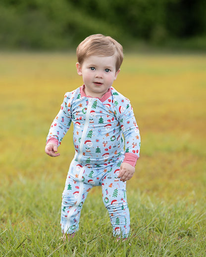 A toddler standing in a grassy field wearing a light blue long-sleeve pajama set with a Christmas tree and Santa pattern, pink ribbed collar and cuffs, and a pink bib-style neckline.