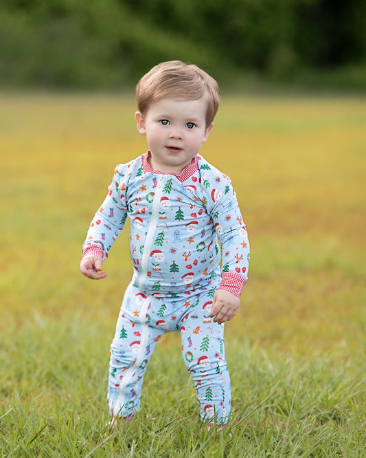 A toddler standing in a grassy field wearing a light blue long-sleeve pajama set with a Christmas tree and Santa pattern, pink ribbed collar and cuffs, and a pink bib-style neckline.