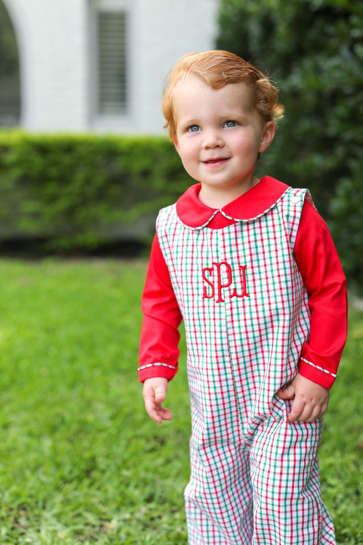 A toddler standing outdoors on grass wearing a red and white gingham overall jumpsuit with a red long-sleeve shirt underneath and red monogram lettering on the chest.